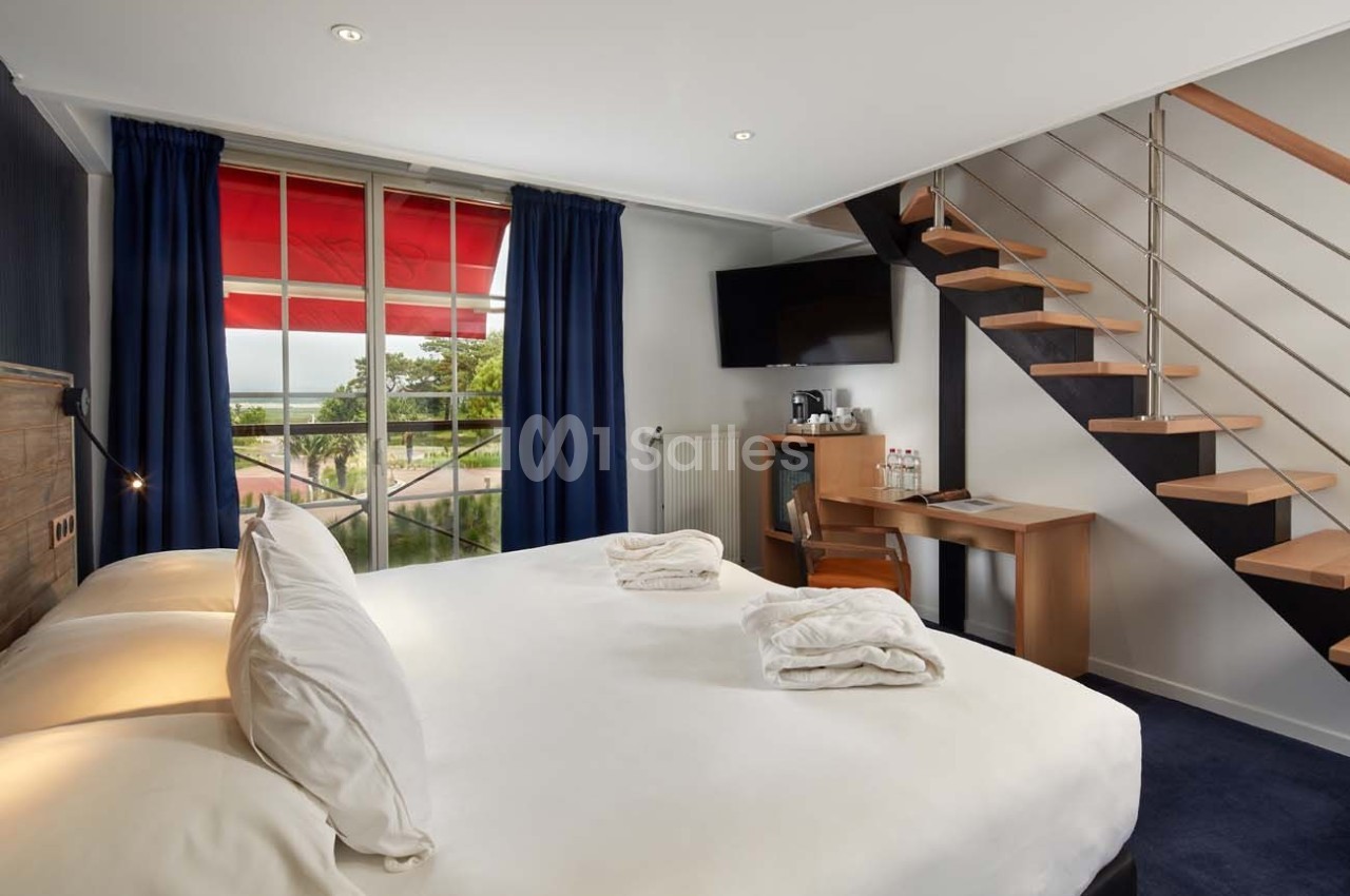 Chambre lumineuse avec lit double, bureau, télévision, escalier en bois et grande fenêtre avec rideaux bleus.