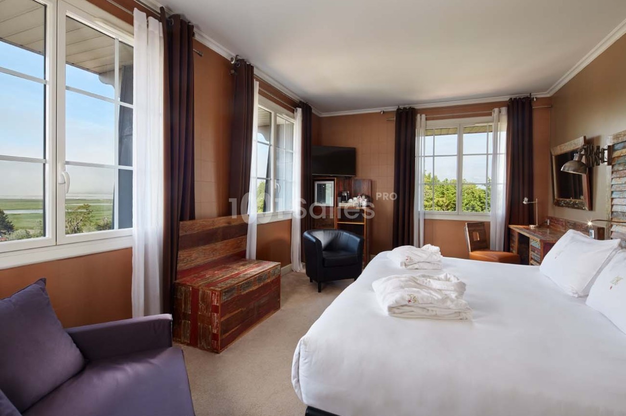 Chambre lumineuse avec lit double, fauteuil, bureau en bois, grandes fenêtres et vue sur un paysage verdoyant.