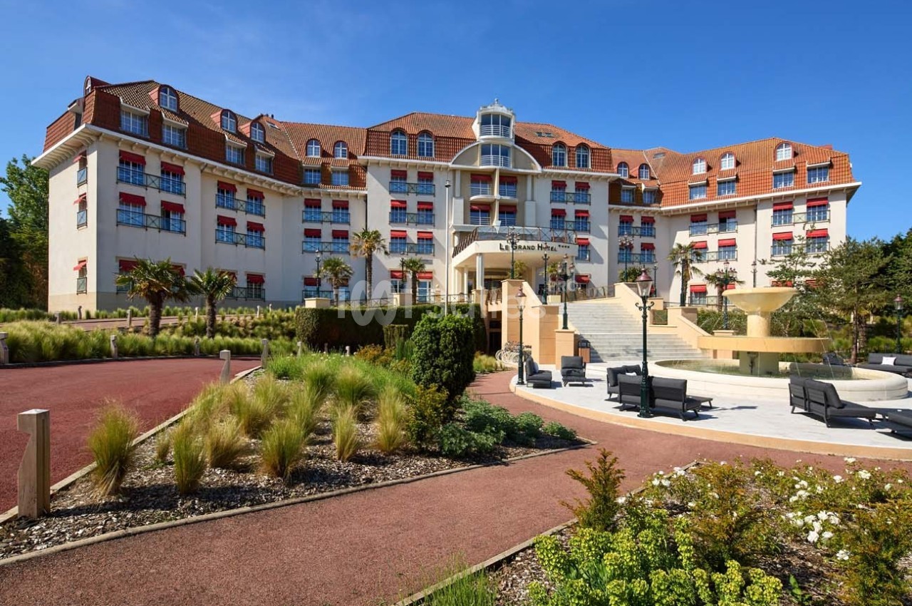 Façade d'un grand hôtel avec toit en tuiles rouges, fenêtres à volets rouges et jardin aménagé au premier plan.