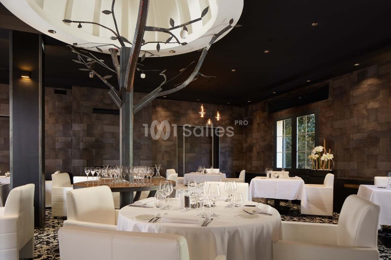 Salle de restaurant élégante avec tables rondes dressées, chaises blanches et décoration moderne incluant un arbre stylisé.