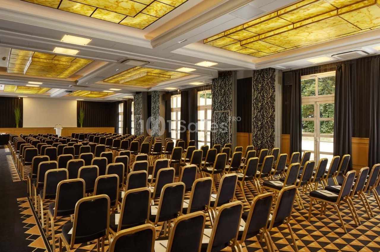 Salle de conférence lumineuse avec rangées de chaises noires, plafond décoratif et grandes fenêtres donnant sur l'extérieur.