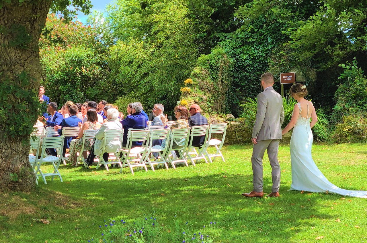 Un couple en tenue de mariage marche vers des invités assis en plein air dans un jardin verdoyant.