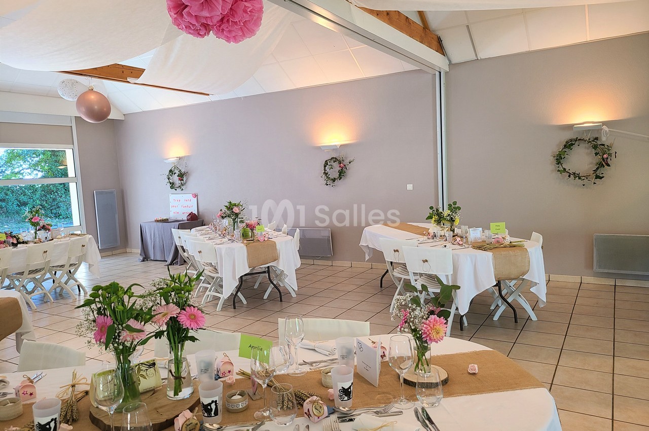 Salle décorée pour une réception avec tables dressées, nappes blanches, fleurs roses et éléments en toile de jute.