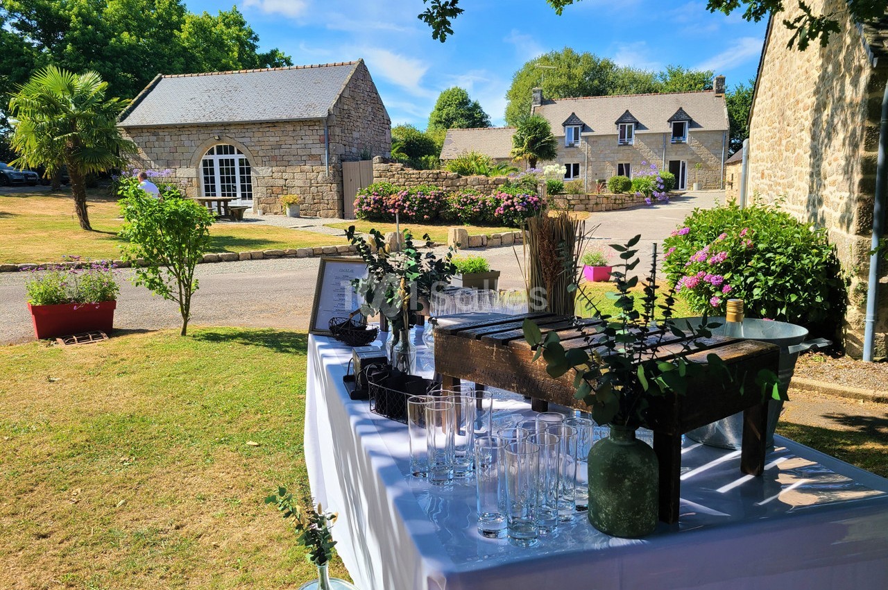 Table dressée en extérieur avec verres et décorations végétales, dans un jardin entouré de bâtiments en pierre.