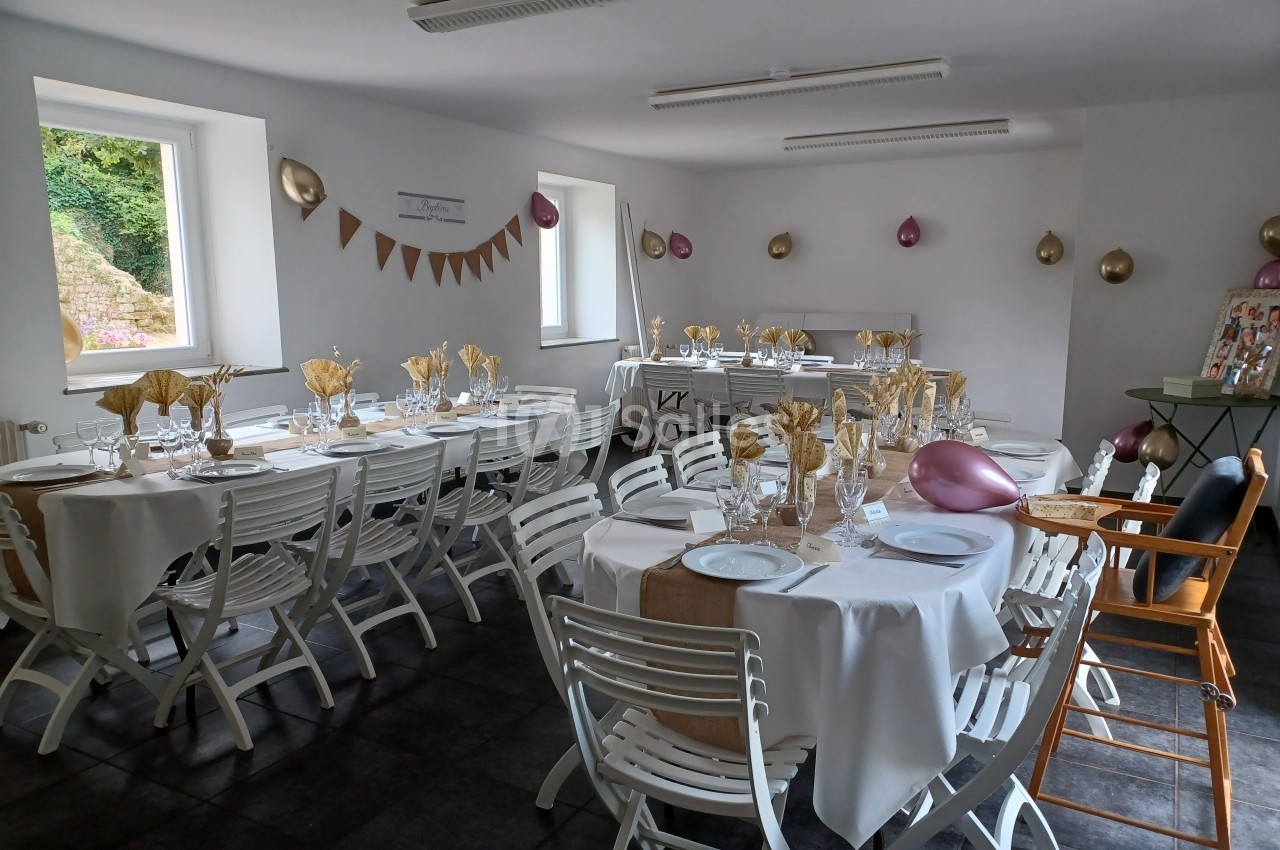 Salle décorée pour un événement avec tables dressées, nappes blanches, chaises pliantes et décorations dorées et roses.