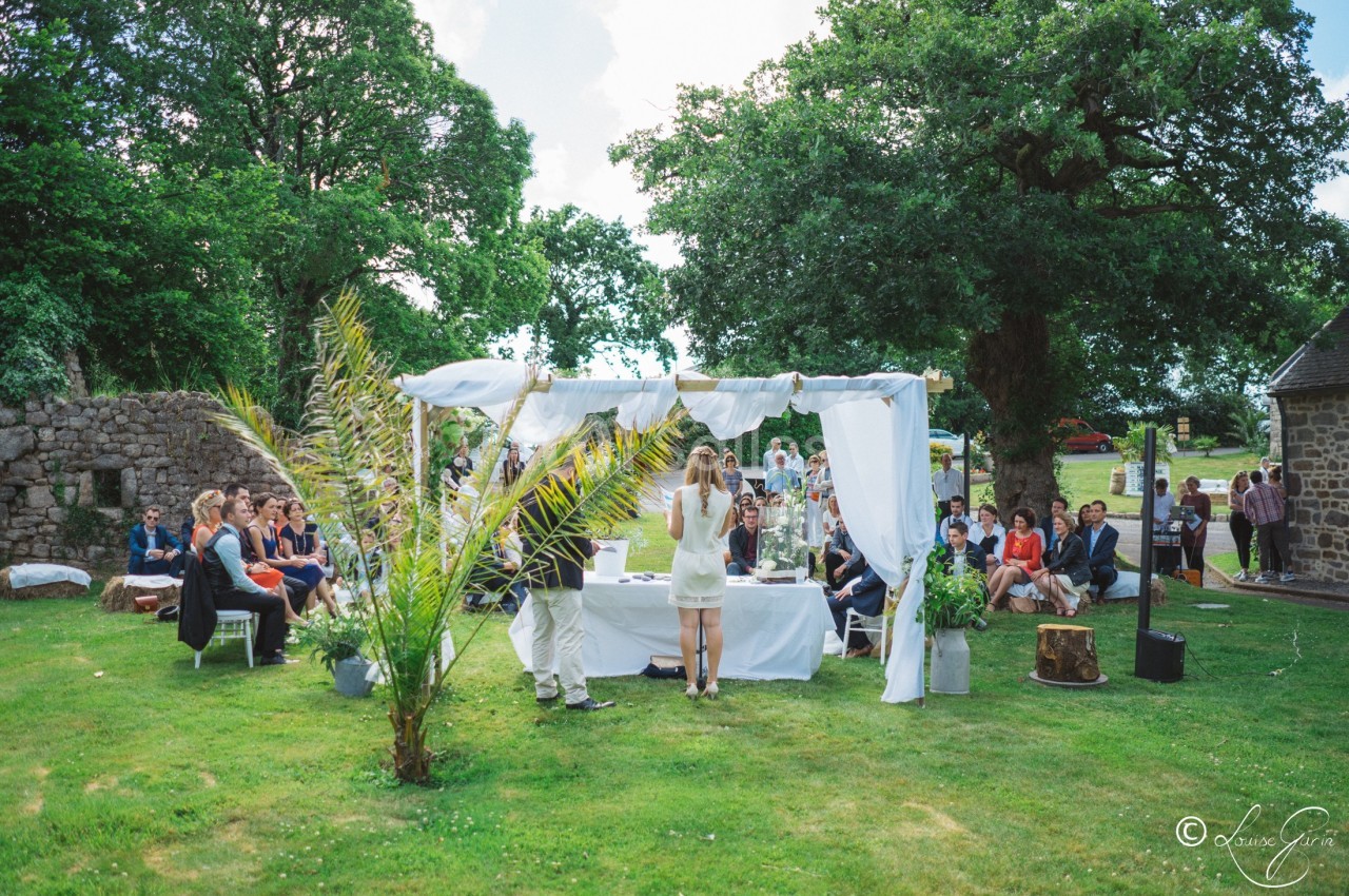 Cérémonie en plein air sous une arche décorée de voilages blancs, entourée d'invités assis sur une pelouse verdoyante.