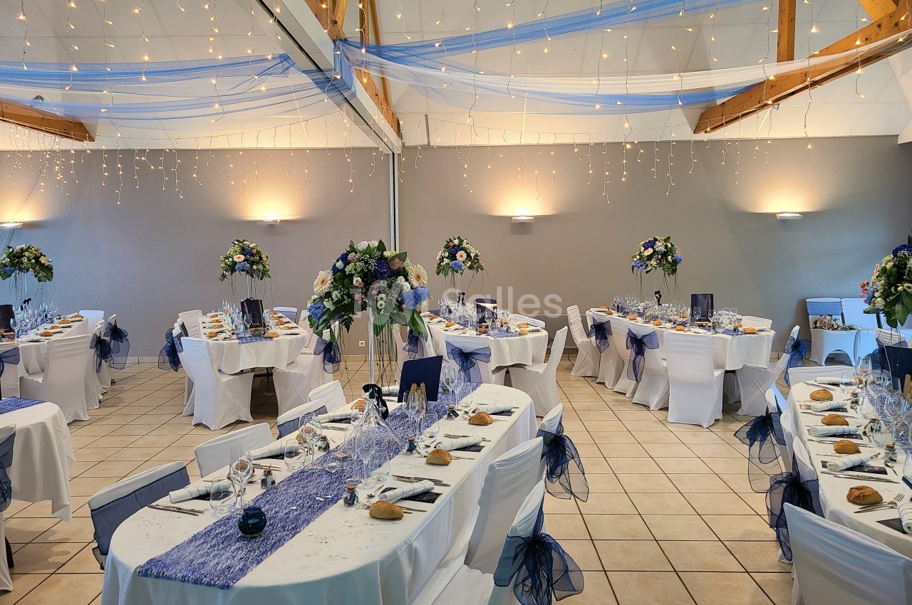 Salle de réception décorée avec des tables dressées, nappes blanches, chemins de table bleus et guirlandes lumineuses…
