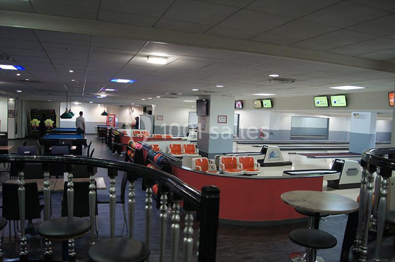 Salle de bowling avec plusieurs pistes, sièges orange, tables hautes et écrans suspendus affichant des scores.