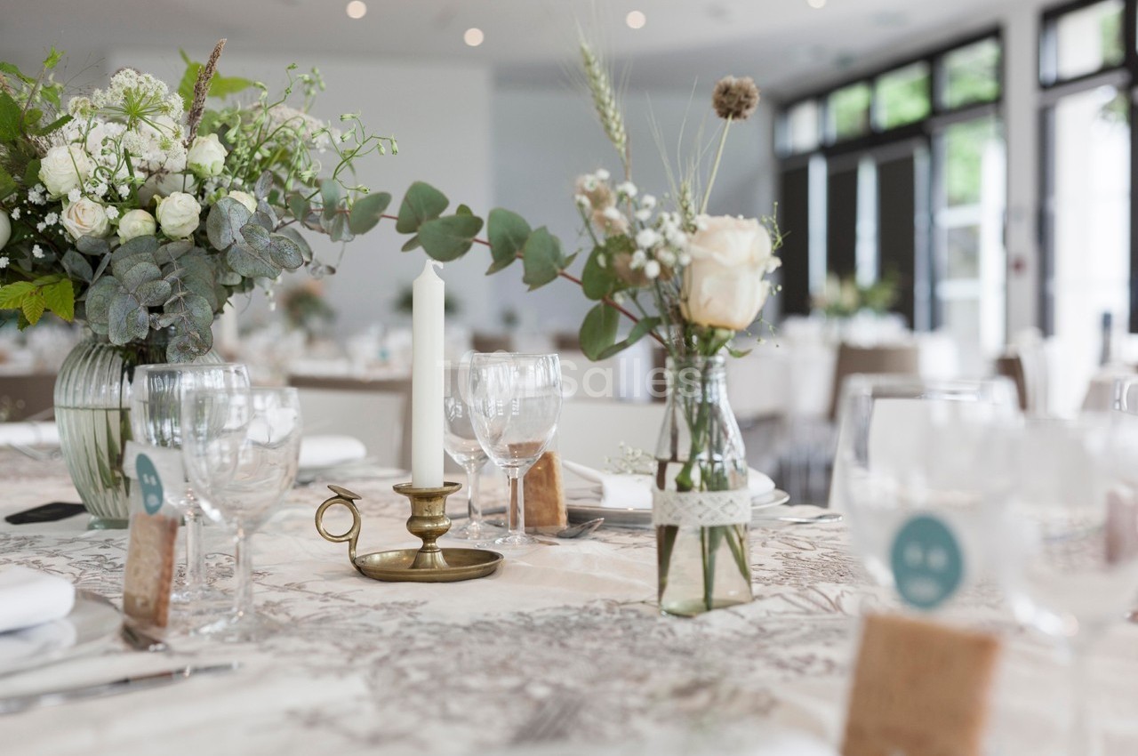 Table décorée avec des fleurs, bougeoir en laiton et vaisselle élégante dans une salle lumineuse.