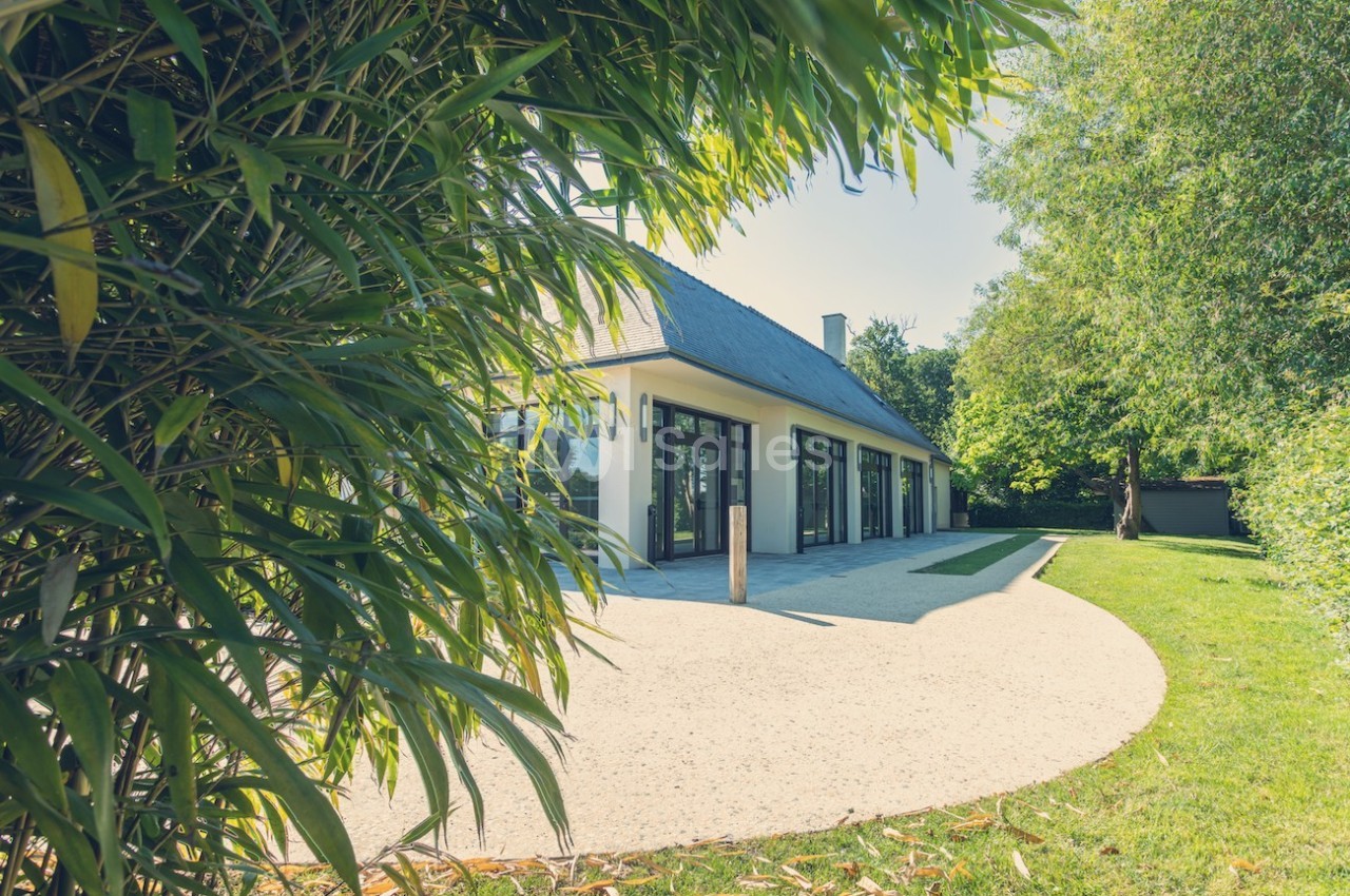 Façade d'une maison moderne avec de grandes baies vitrées, entourée de végétation et d'une allée en gravier.