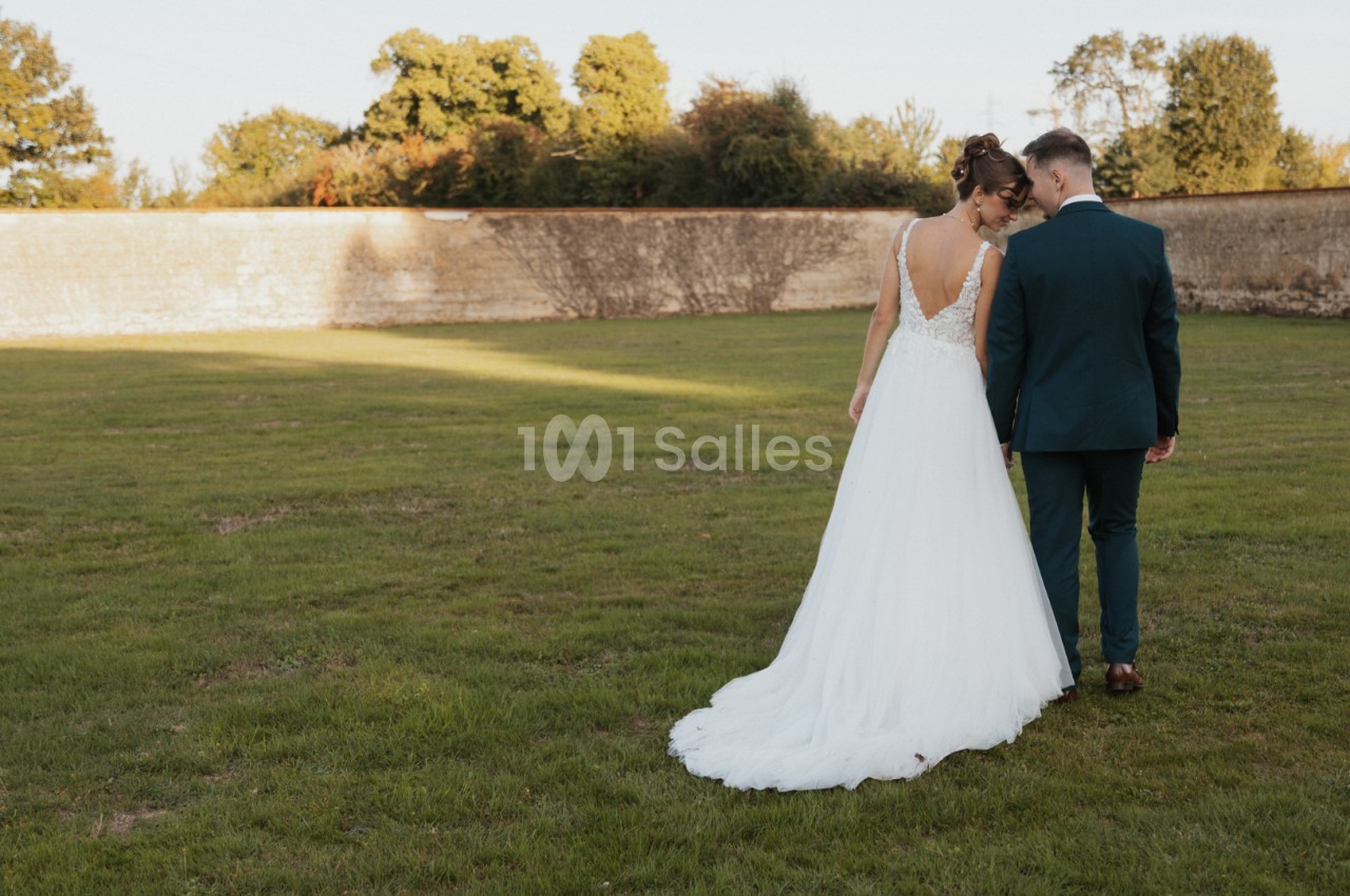 Une mariée en robe blanche et un marié en costume vert marchent côte à côte dans un jardin.