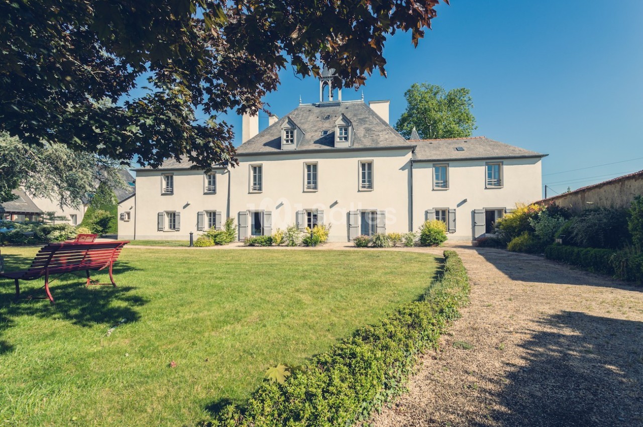 Grande maison traditionnelle avec façade blanche, entourée d'un jardin verdoyant et d'une allée gravillonnée.