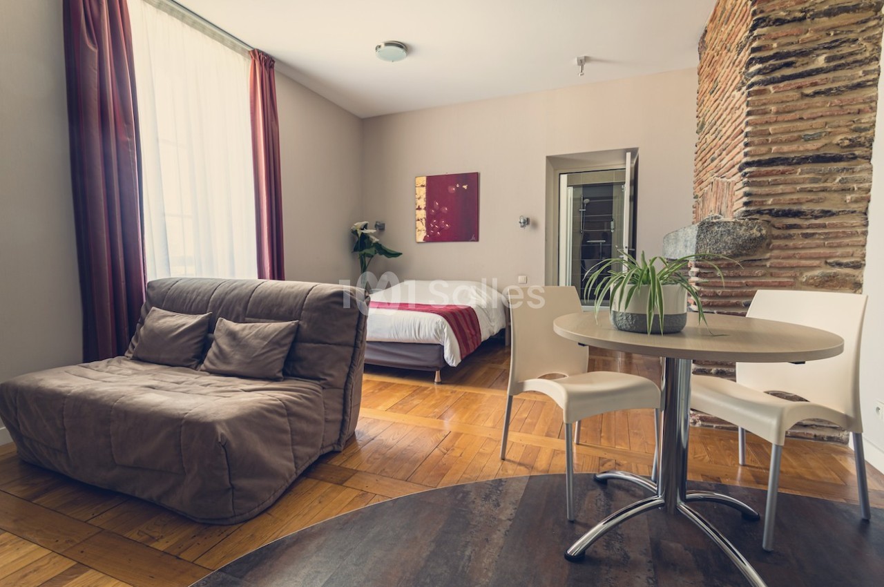 Studio lumineux avec parquet, canapé, table ronde, chaises blanches, lit double et mur en briques apparentes.