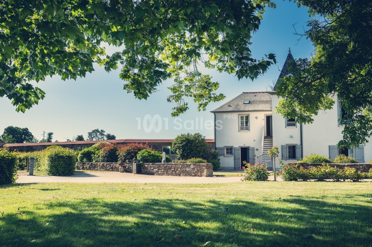 Maison blanche traditionnelle avec jardin verdoyant, arbres ombragés et ciel bleu en arrière-plan.