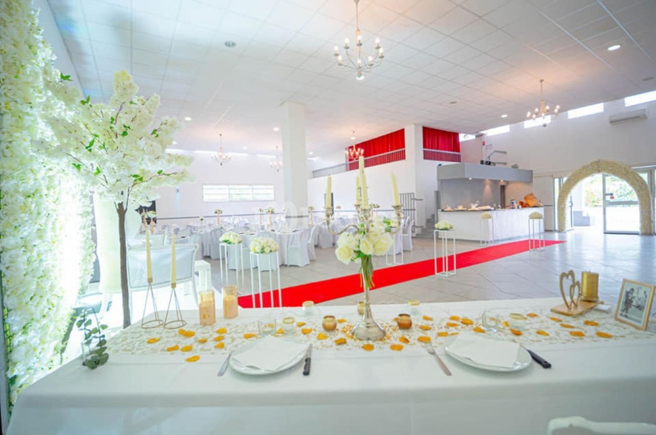Salle de réception lumineuse décorée pour un mariage avec une table d'honneur ornée de fleurs et bougies.