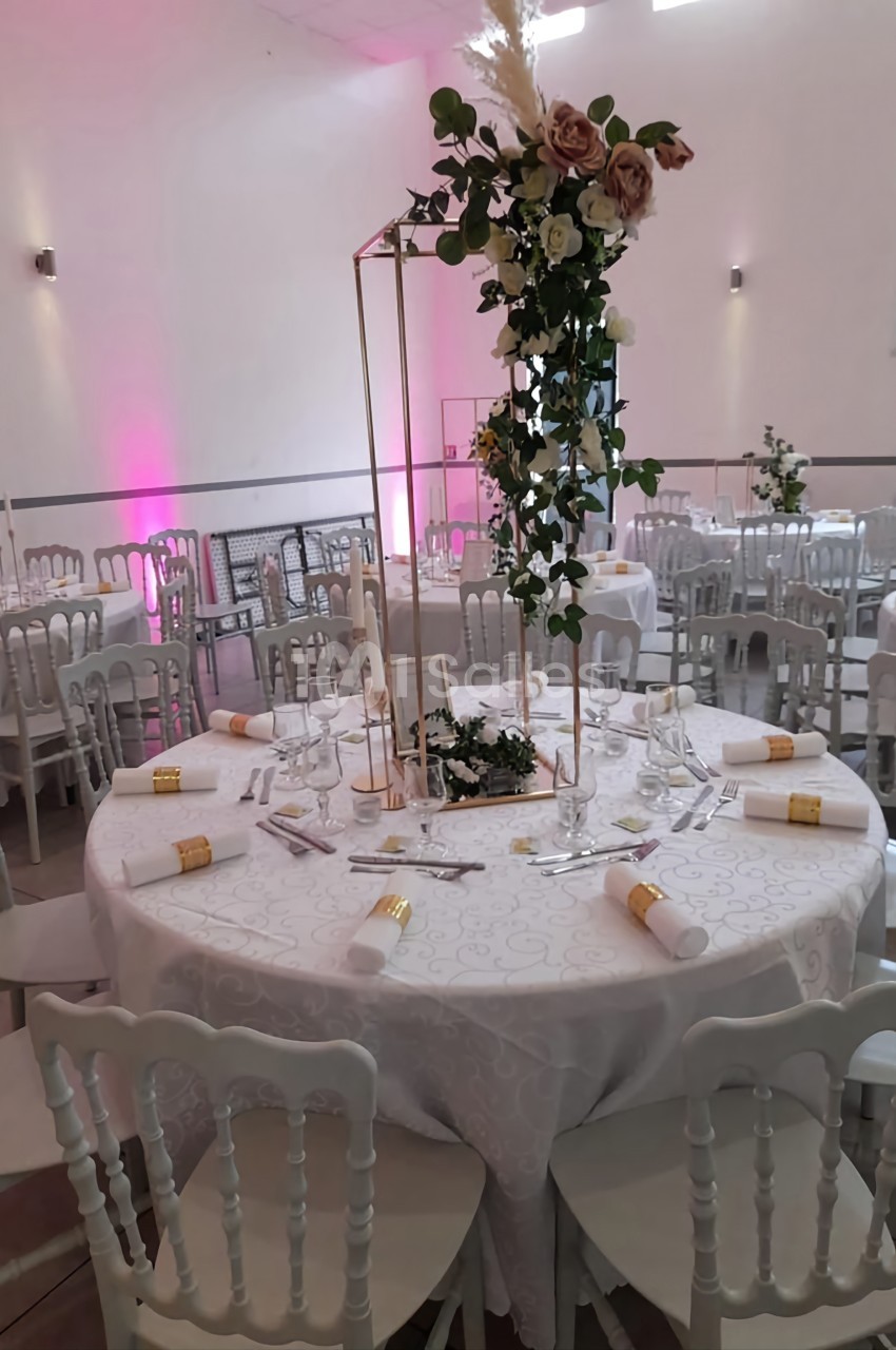 Salle de réception décorée avec des tables rondes, nappes blanches, chaises élégantes et centres de table fleuris.