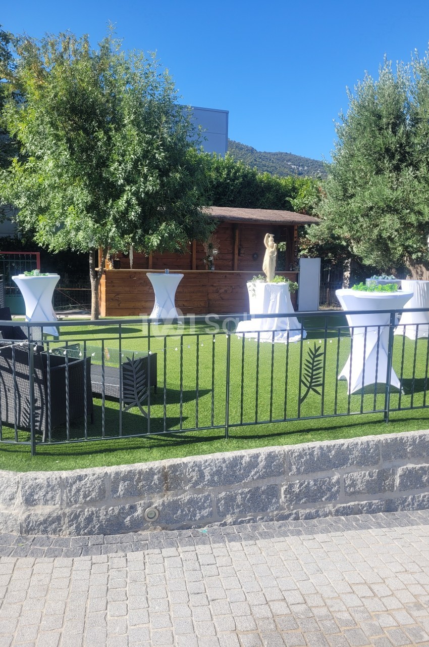 Espace extérieur aménagé avec pelouse artificielle, tables hautes, fauteuils en rotin et cabanon en bois, entouré de verdure.