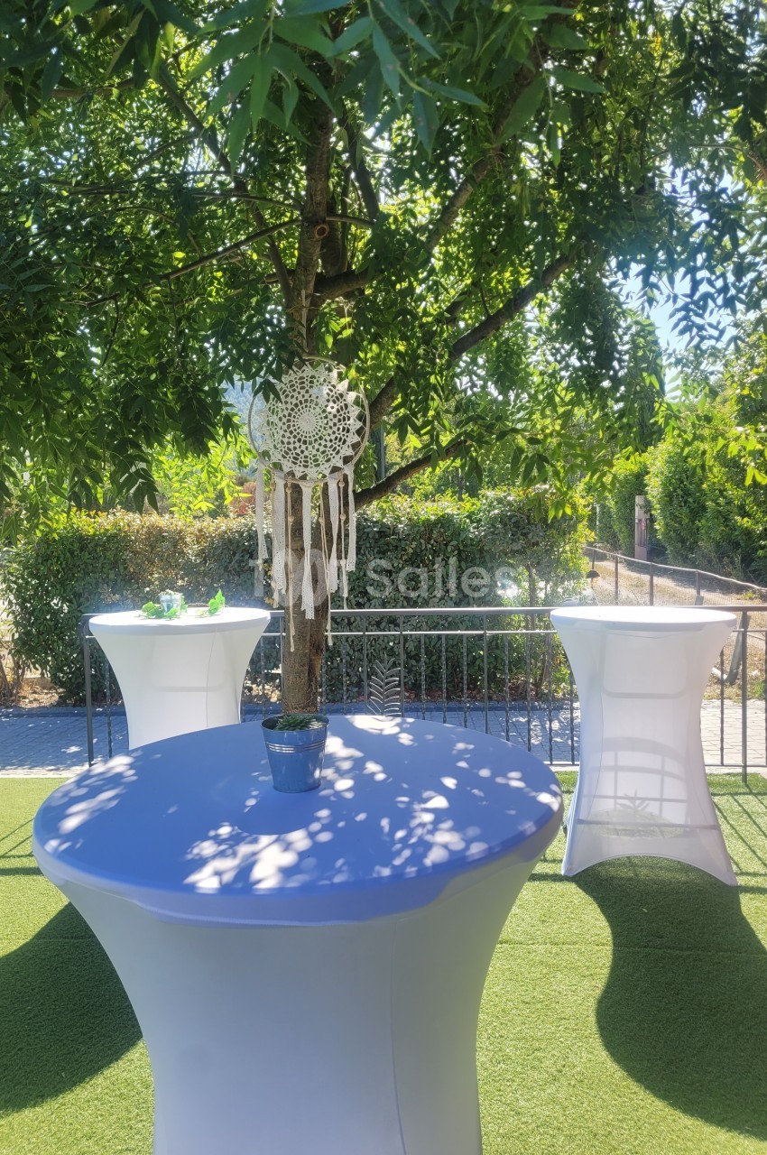 Table haute habillée de blanc sous un arbre, décorée d'un attrape-rêves et entourée de verdure en extérieur.