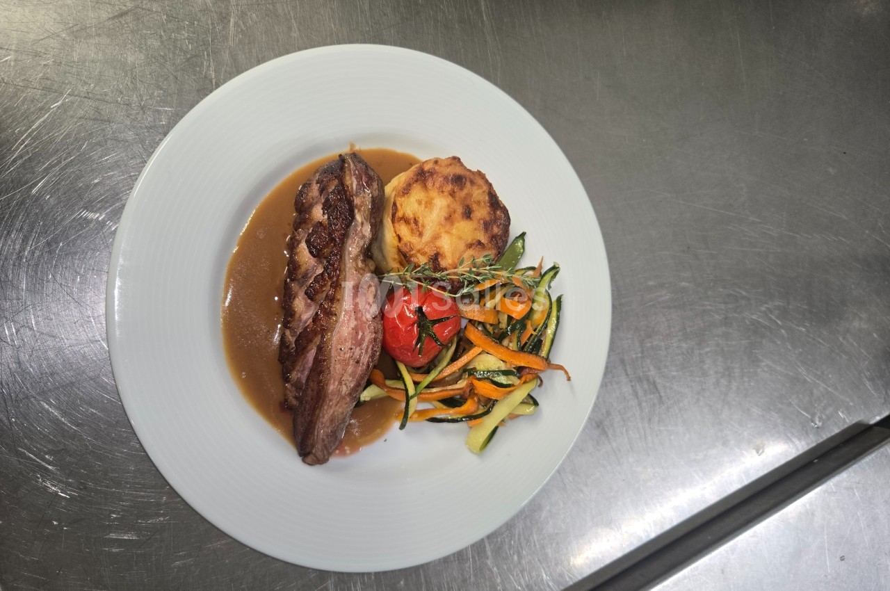 Assiette contenant une pièce de viande avec sauce, gratin de pommes de terre et légumes variés sur une table en inox.