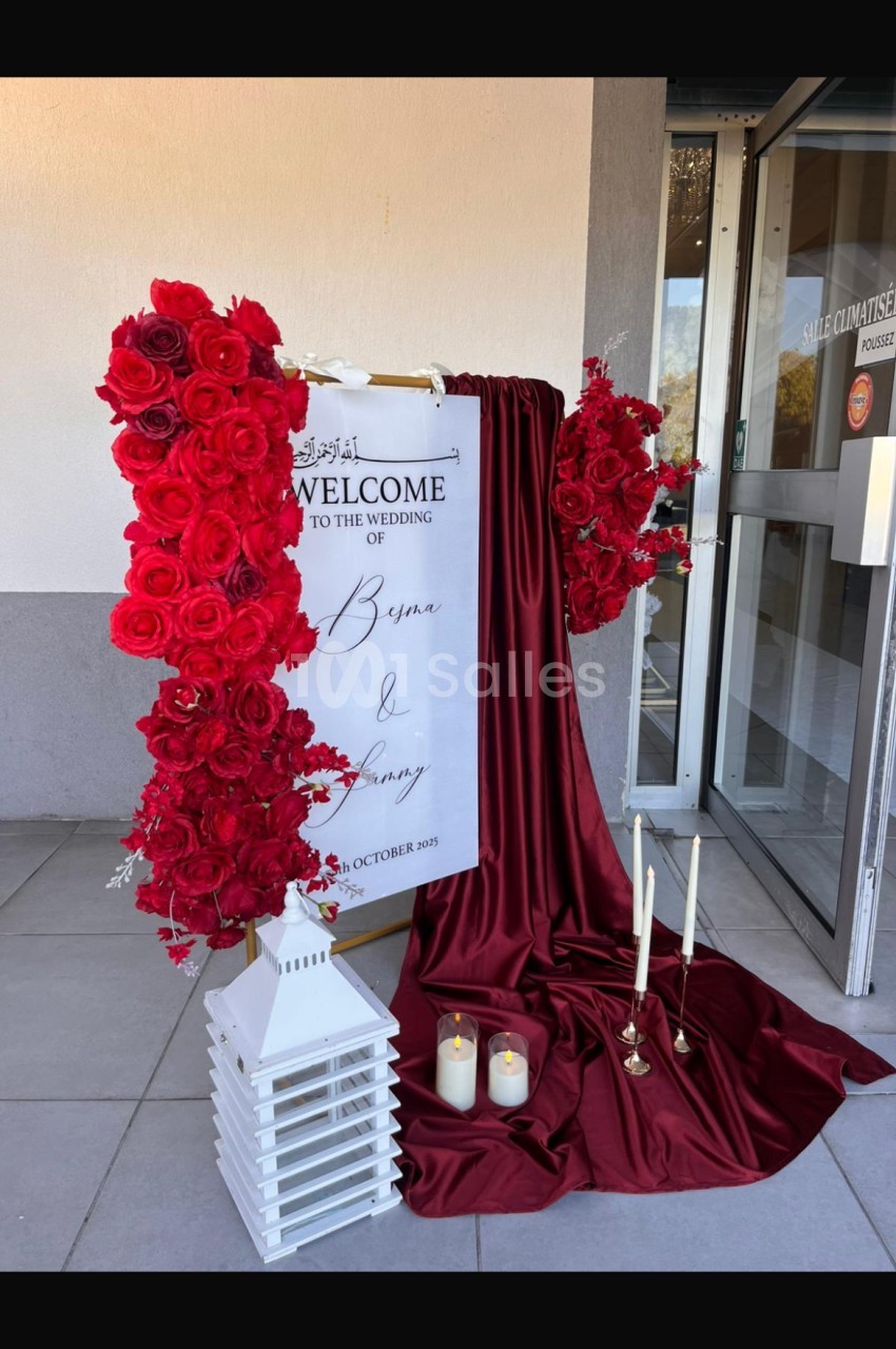 Panneau de bienvenue pour un mariage entouré de roses rouges, drapé bordeaux, bougies et lanterne blanche.