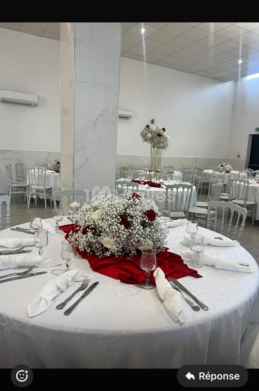 Salle de réception avec tables rondes décorées de nappes blanches, centres de table floraux rouges et blancs.