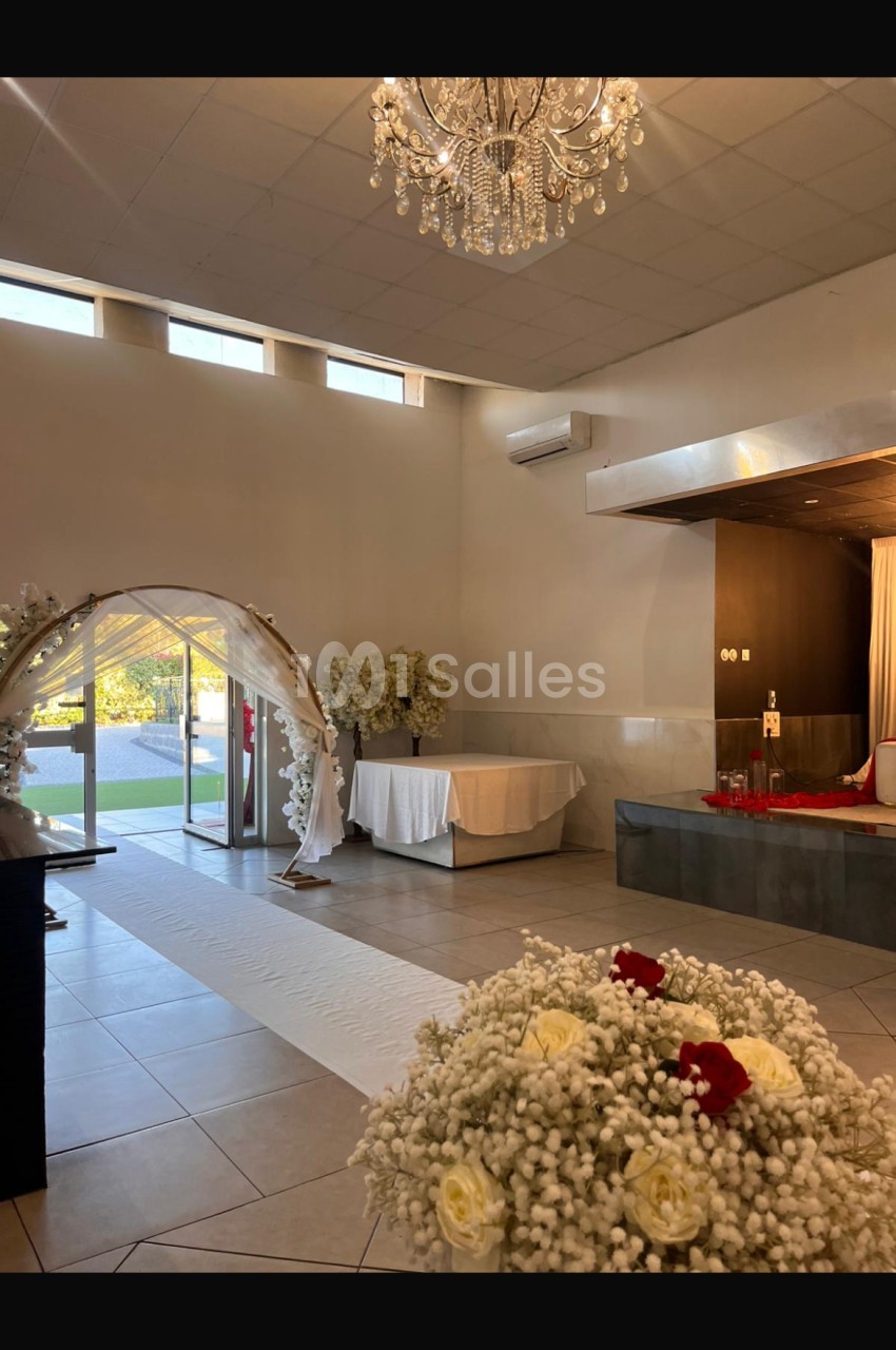 Salle de réception lumineuse avec arche décorative, fleurs blanches, lustre et table dressée près d'une baie vitrée.