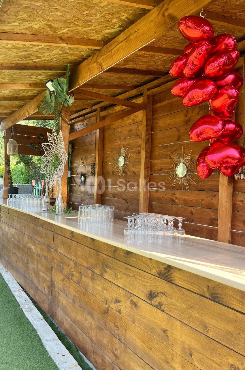 Comptoir en bois décoré de ballons rouges en forme de cœur et de verres alignés, situé en extérieur sous un auvent.