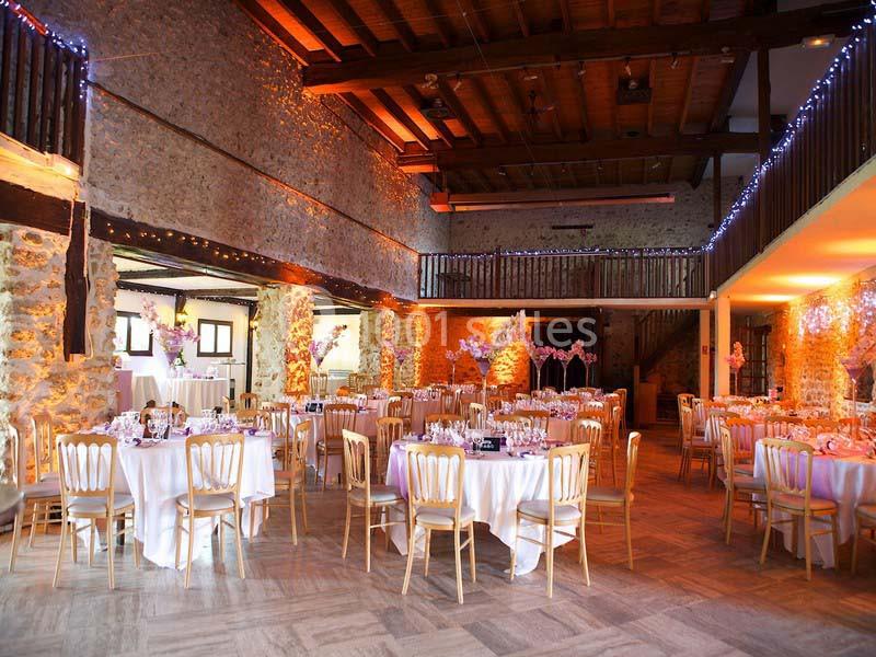Salle de réception en pierre avec tables rondes dressées, chaises en bois clair et guirlandes lumineuses suspendues.