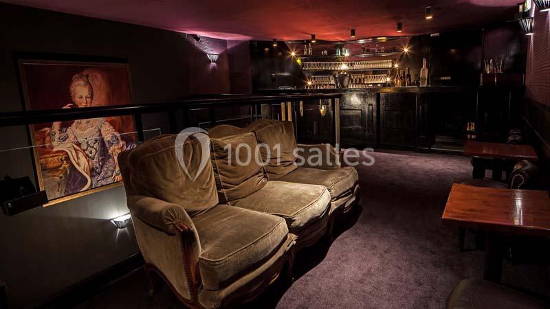 Salon cosy avec fauteuils en velours, table en bois et bar éclairé en arrière-plan dans une ambiance tamisée.