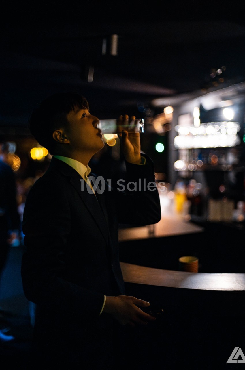 Personne en costume buvant un verre dans un bar faiblement éclairé, avec des lumières floues en arrière-plan.