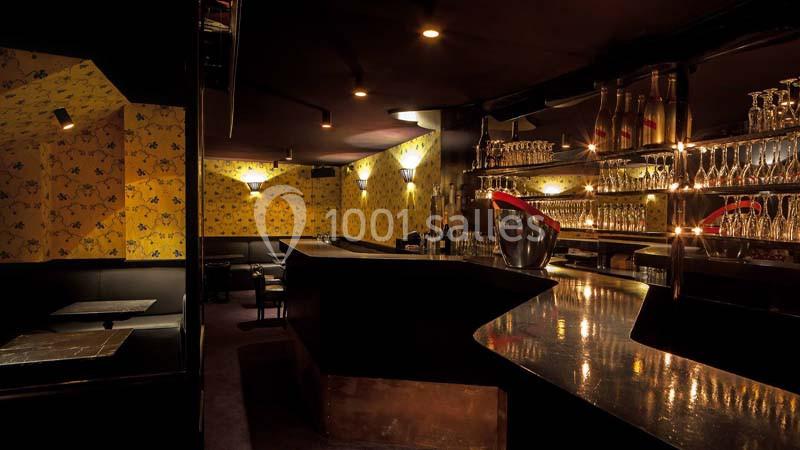 Bar sombre avec comptoir en bois brillant, étagères remplies de verres et bouteilles, banquettes noires en arrière-plan.