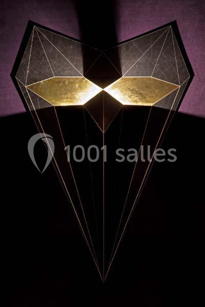 Structure géométrique en forme de masque avec des reflets dorés, éclairée sur un fond sombre.