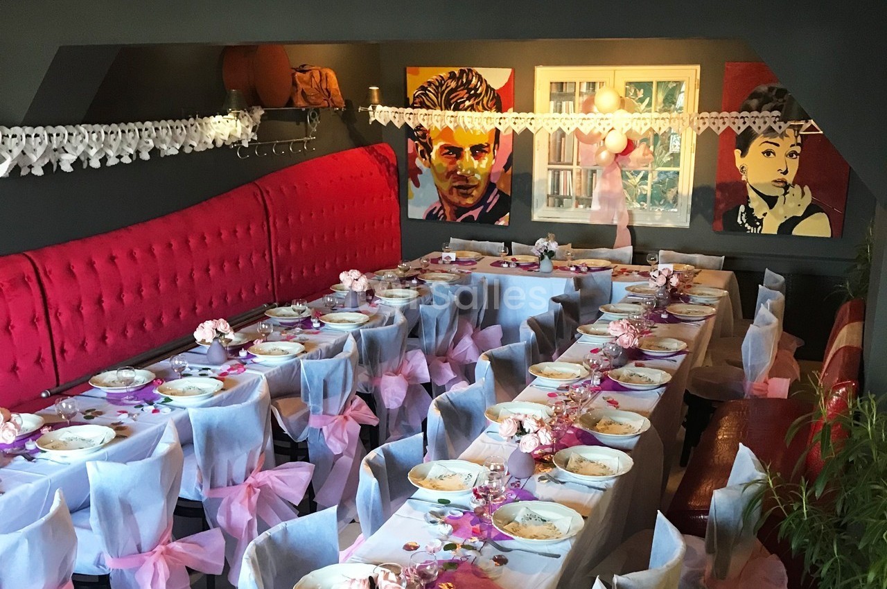 Salle décorée pour un événement, avec tables alignées, nappes blanches, nœuds roses et portraits accrochés au mur.
