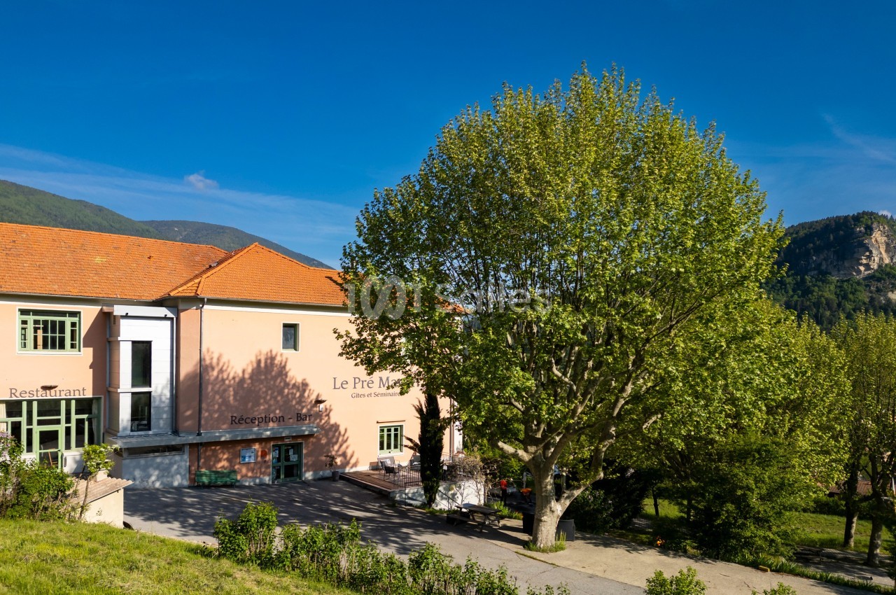 Bâtiment avec façade rose, entouré de verdure, sous un ciel bleu, avec un grand arbre au premier plan.