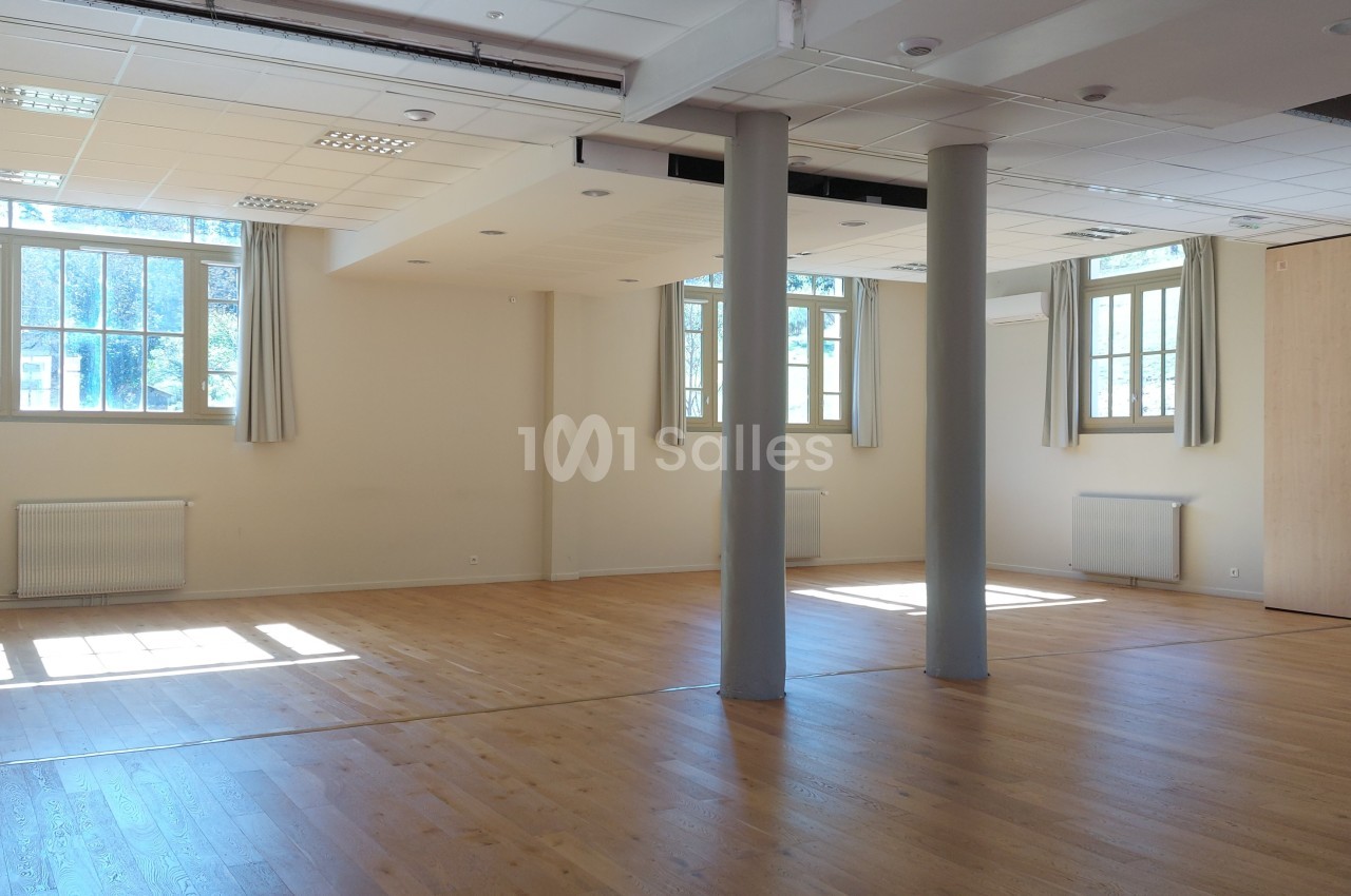 Salle lumineuse avec parquet en bois, grandes fenêtres laissant entrer la lumière naturelle et colonnes au centre.
