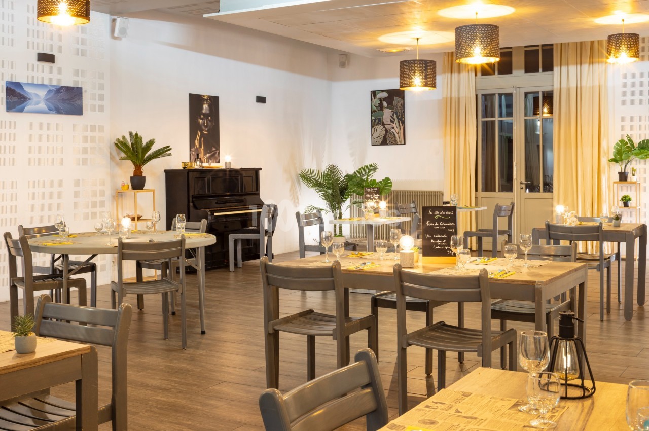 Salle de restaurant lumineuse avec tables en bois, chaises grises, piano noir et décorations végétales.