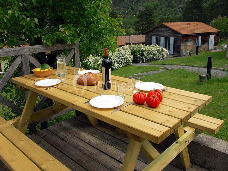 Table de pique-nique en bois dressée avec des assiettes, des tomates, du pain et une bouteille, dans un jardin verdoyant.