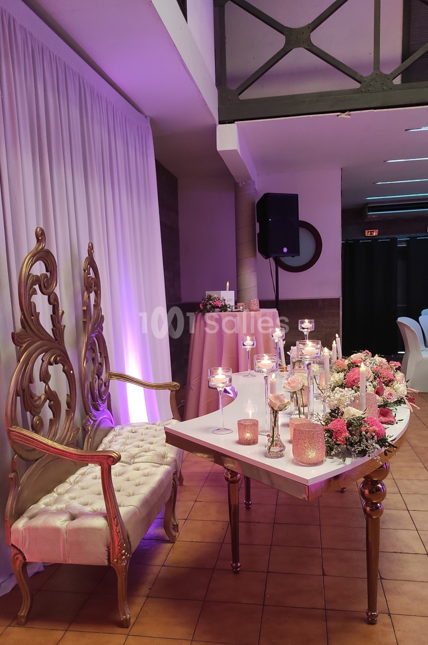 Chaises dorées ornées et table décorée de bougies et fleurs dans une salle de réception aux tons roses et blancs.