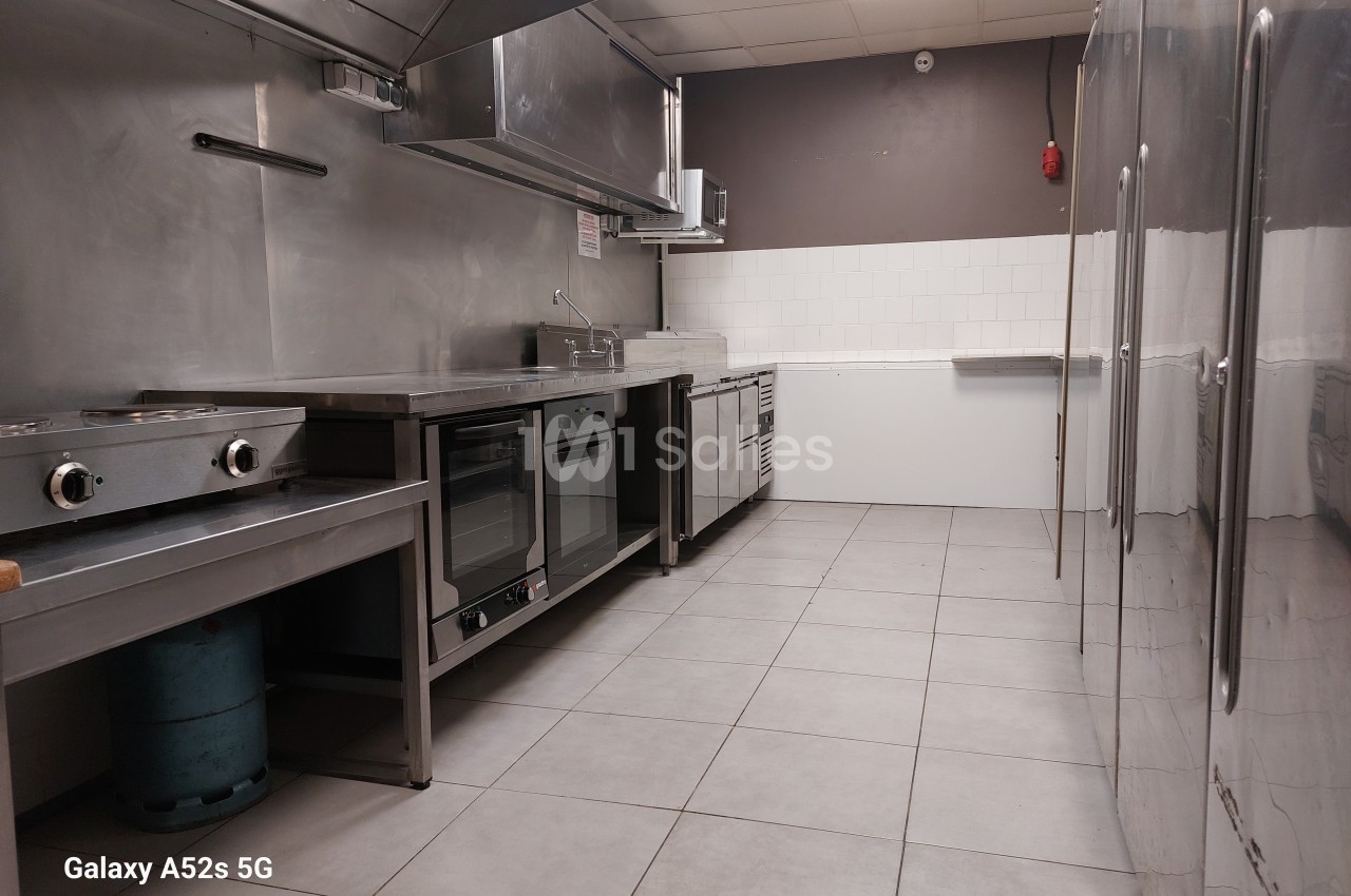 Cuisine professionnelle équipée avec plans de travail en inox, four, plaques de cuisson et carrelage au sol.