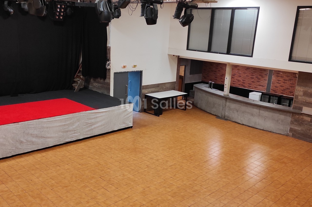Salle polyvalente avec scène surélevée, rideaux noirs, projecteurs, comptoir en angle et sol en carrelage.