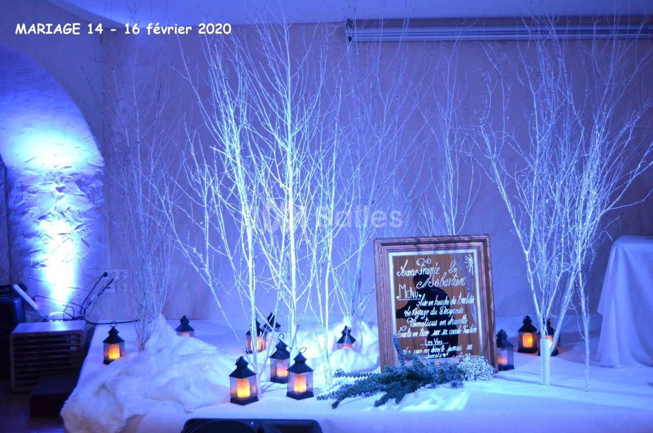 Décoration de mariage hivernale avec branches blanches, lanternes allumées et panneau en bois dans une ambiance bleutée.