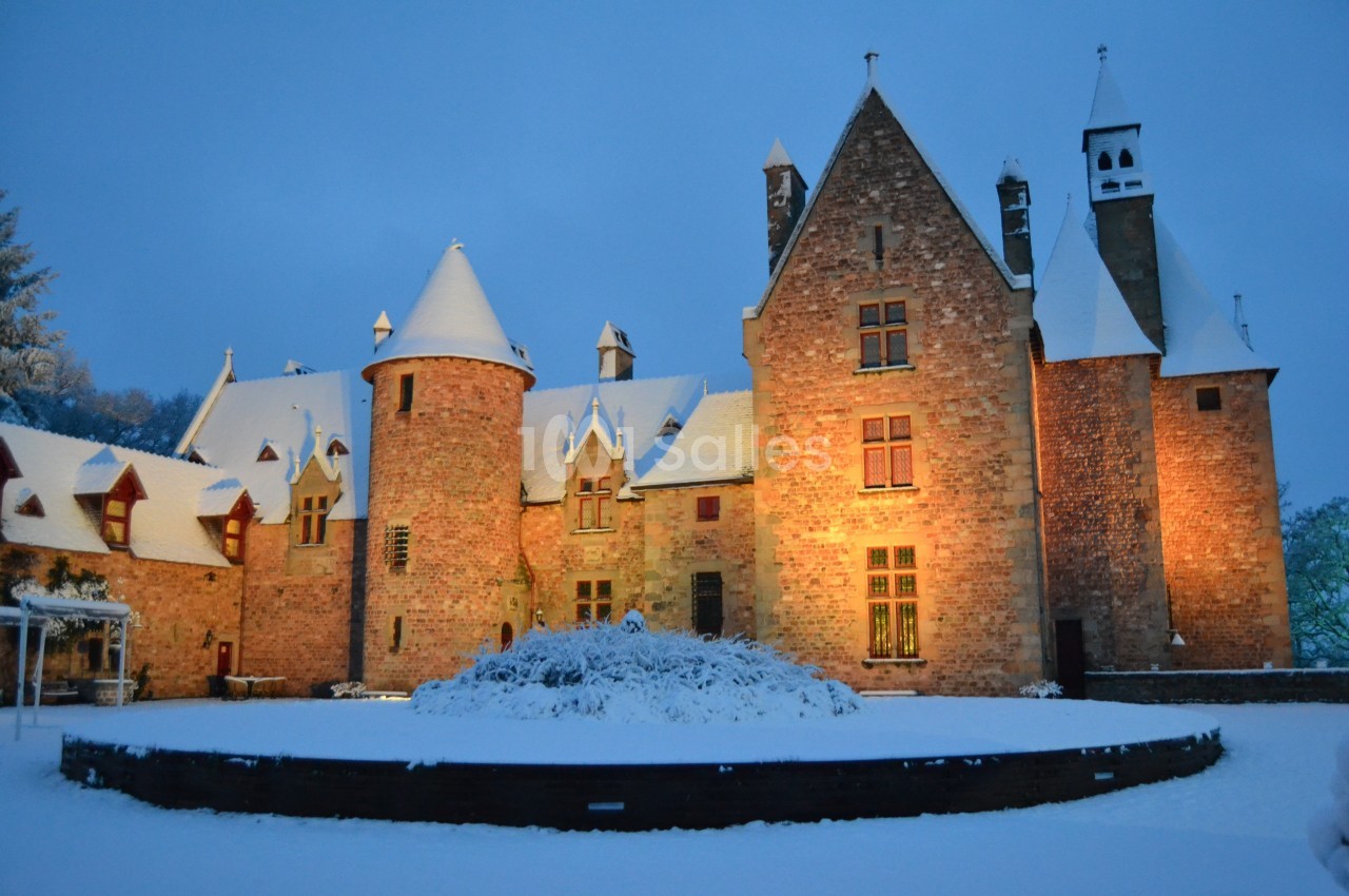 Château en pierre éclairé au crépuscule, entouré de neige et d'arbres sous un ciel hivernal.