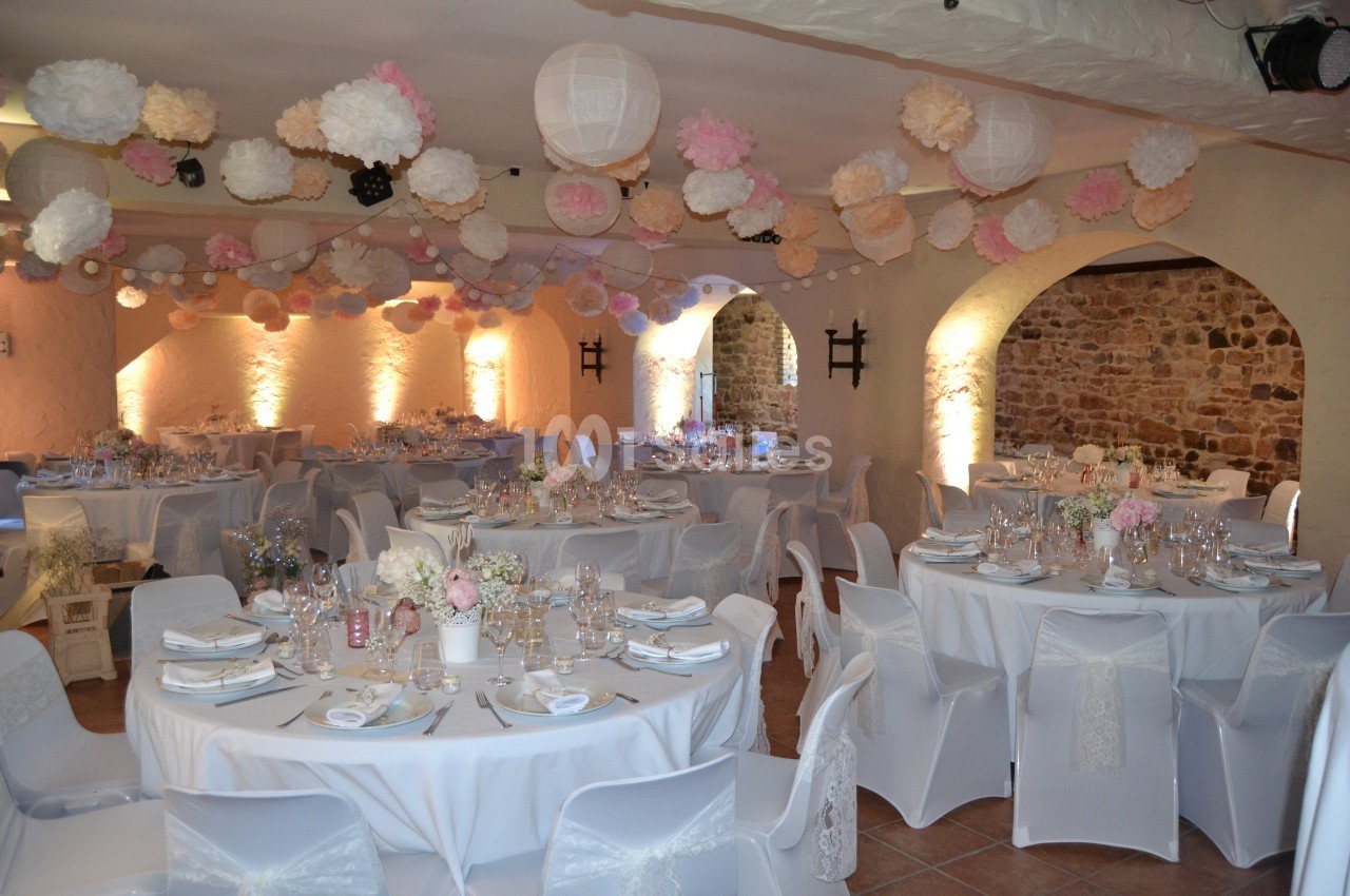 Salle décorée pour un mariage avec tables rondes, nappes blanches, vaisselle élégante et guirlandes de lanternes et pompons…