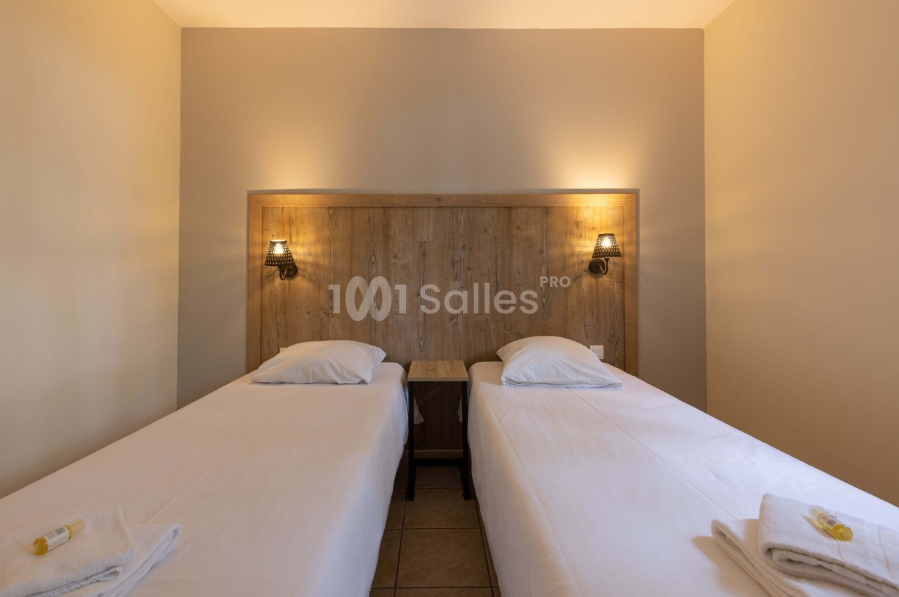 Deux lits simples avec draps blancs, séparés par une table de chevet en bois, éclairés par deux appliques murales.