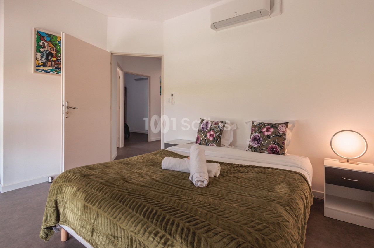 Chambre lumineuse avec lit double, couvre-lit vert, coussins fleuris, table de chevet et porte ouverte sur un couloir.