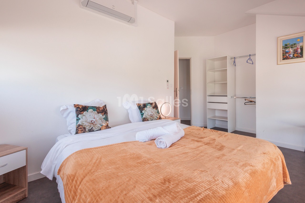 Chambre lumineuse avec lit double, couverture orange, coussins fleuris, placard ouvert et climatisation murale.