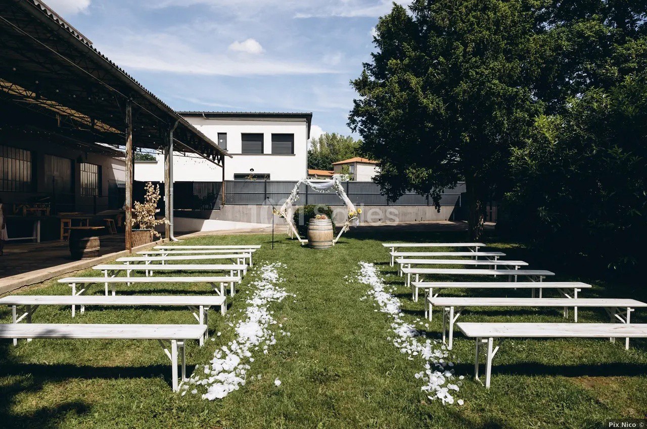 Allée centrale décorée de pétales blancs, entourée de bancs en bois, menant à une arche pour une cérémonie en extérieur.