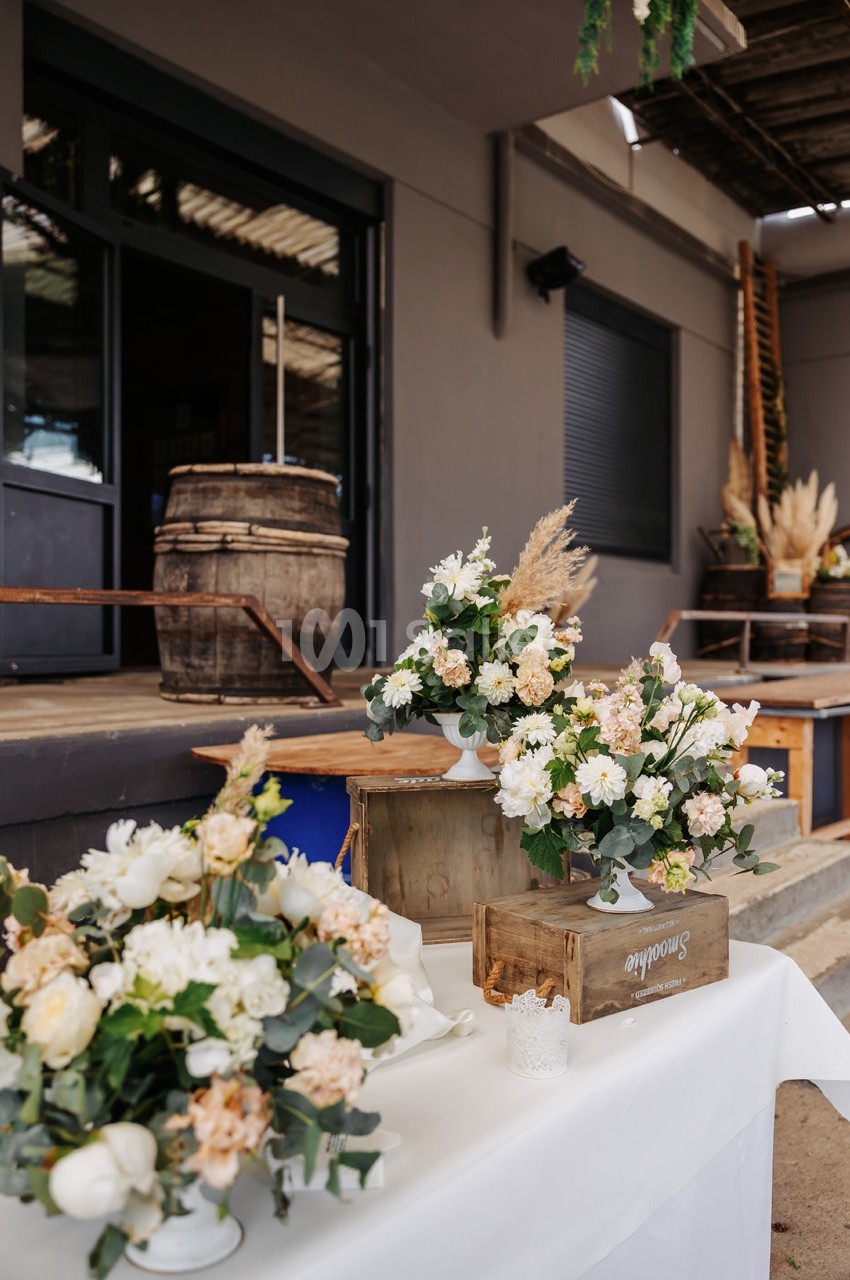 Arrangements floraux blancs et beiges sur une table en extérieur, près de caisses en bois et d'un bâtiment industriel.