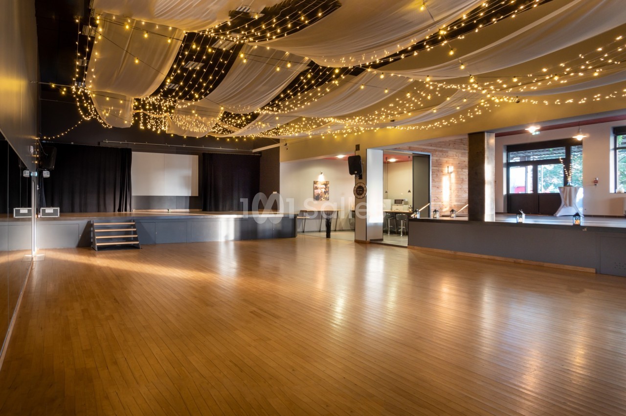 Salle spacieuse avec parquet, scène, rideaux noirs et plafond décoré de guirlandes lumineuses et de drapés blancs.