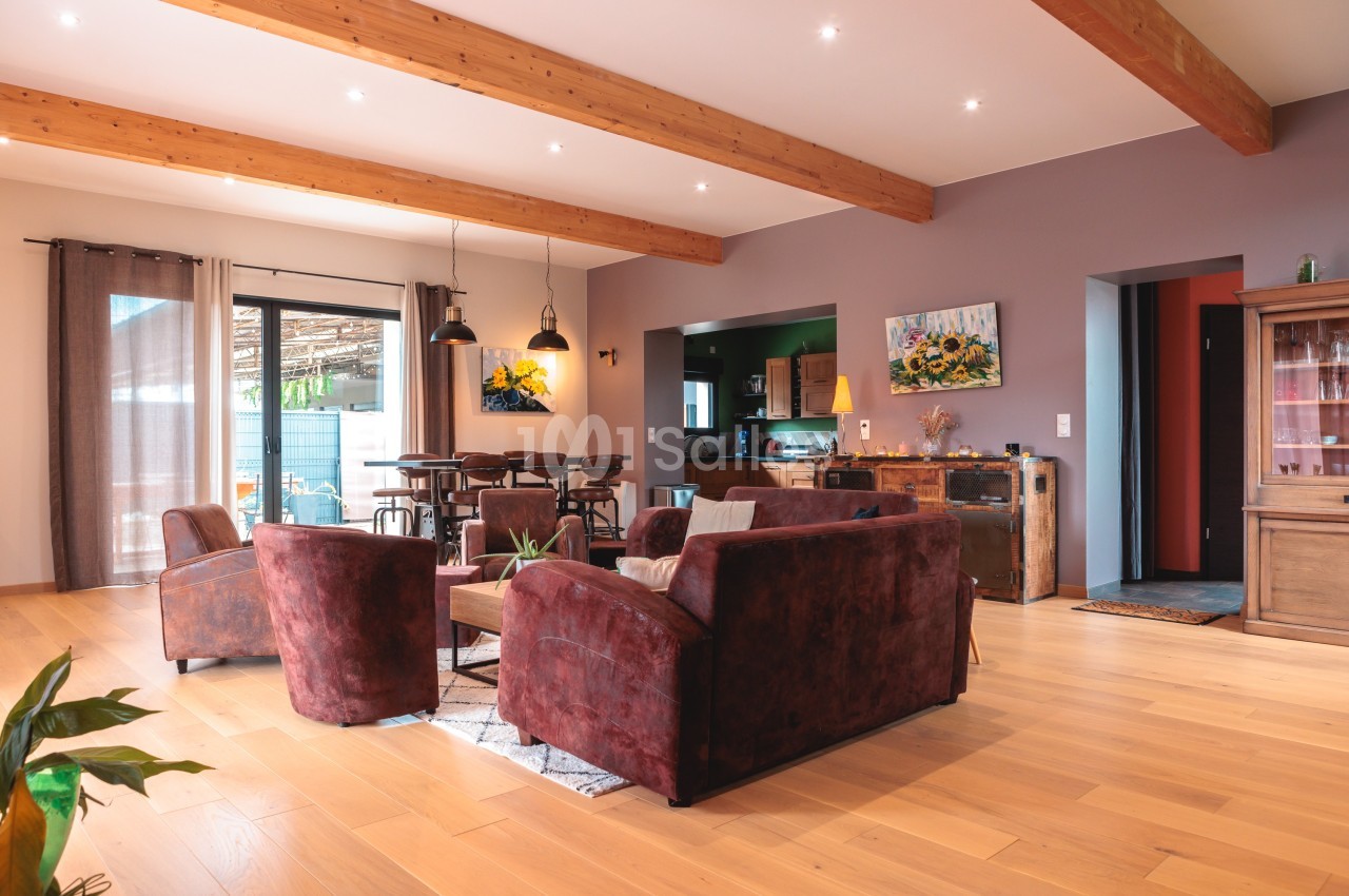 Salon spacieux avec parquet clair, canapés en velours marron, poutres apparentes et lumière naturelle abondante.